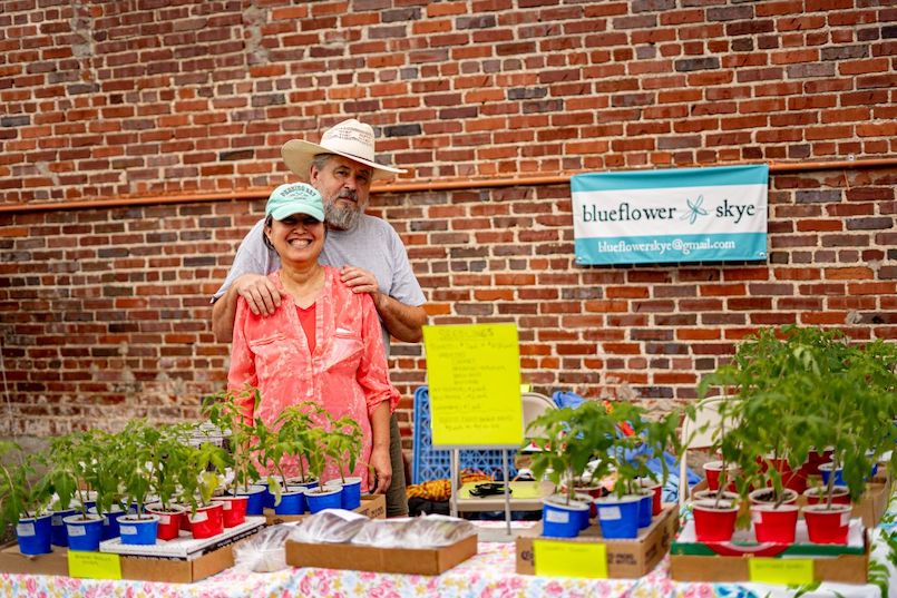 The Blueflower Skye booth at the farmers market. The owners of the booth are smiling from behind a booth filled with solo cups holding small plants.