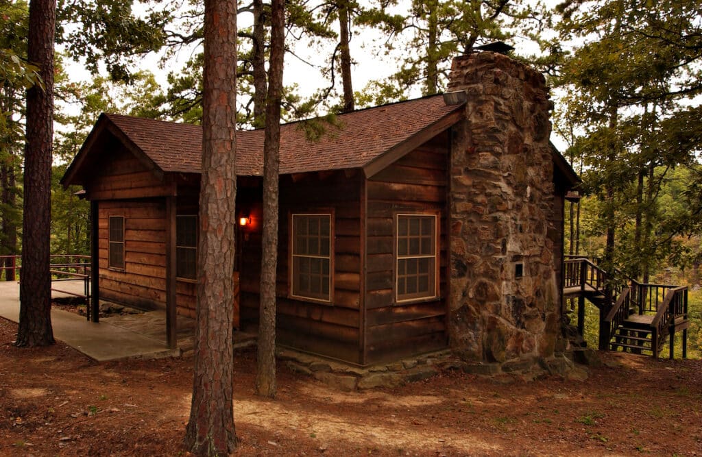 A wooden cabin at Petit Jean State Park. The cabin is made of dark wood and has a large stone chimney on the right side. Steep stairs can be seen leading down from the back deck of the cabin. There are trees surrounding the cabin and a gas lamp by the front door.