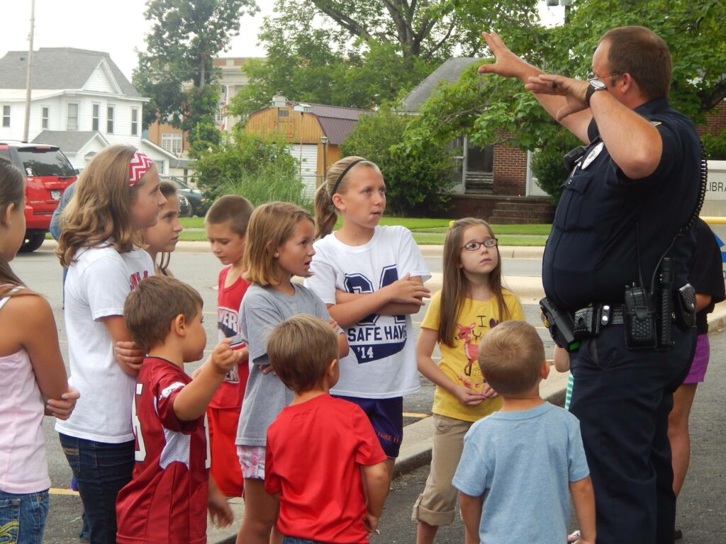 A police officer giving direction to a group of young kids dressed in outdoor clothes for summer.