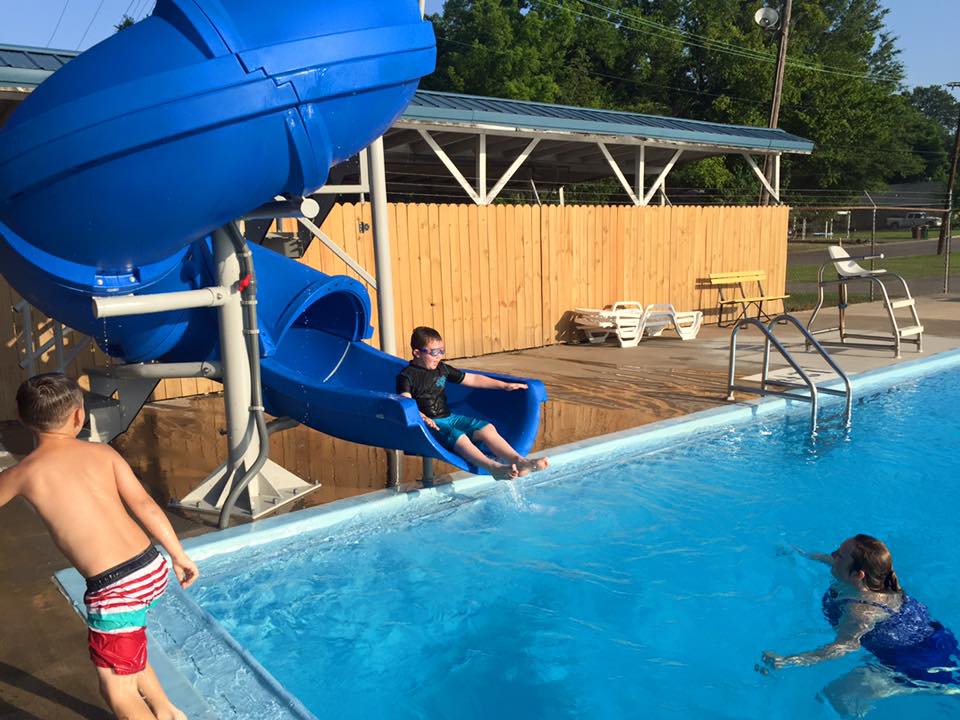 Kids going down a blue swirly slide at a pool. The boys mom is at the bottom of the pool to catch him.
