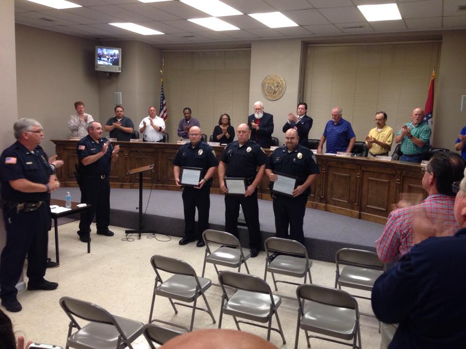 Police Officers receiving framed certificates. A board of people are standing behind them clapping and an audience is also giving a standing ovation.