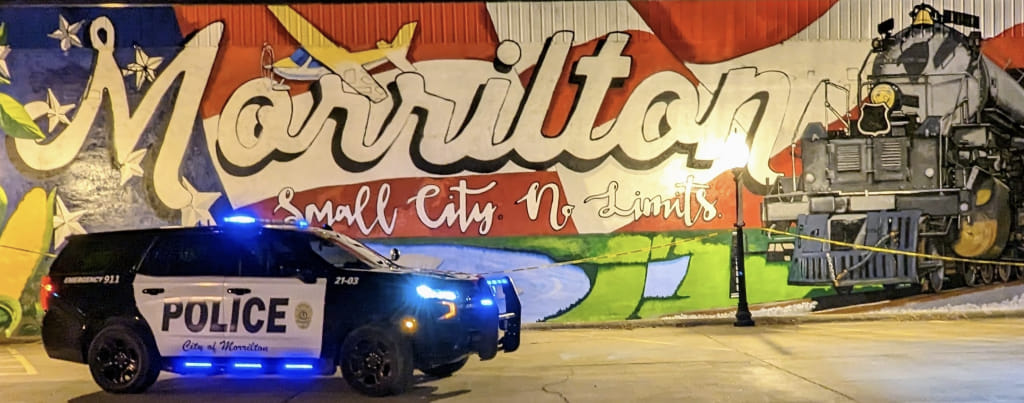 A police car with its lights on sitting in front of the Morrilton wall mural. The mural has an American flag background, a train, a plane, and the bottom is a pond.