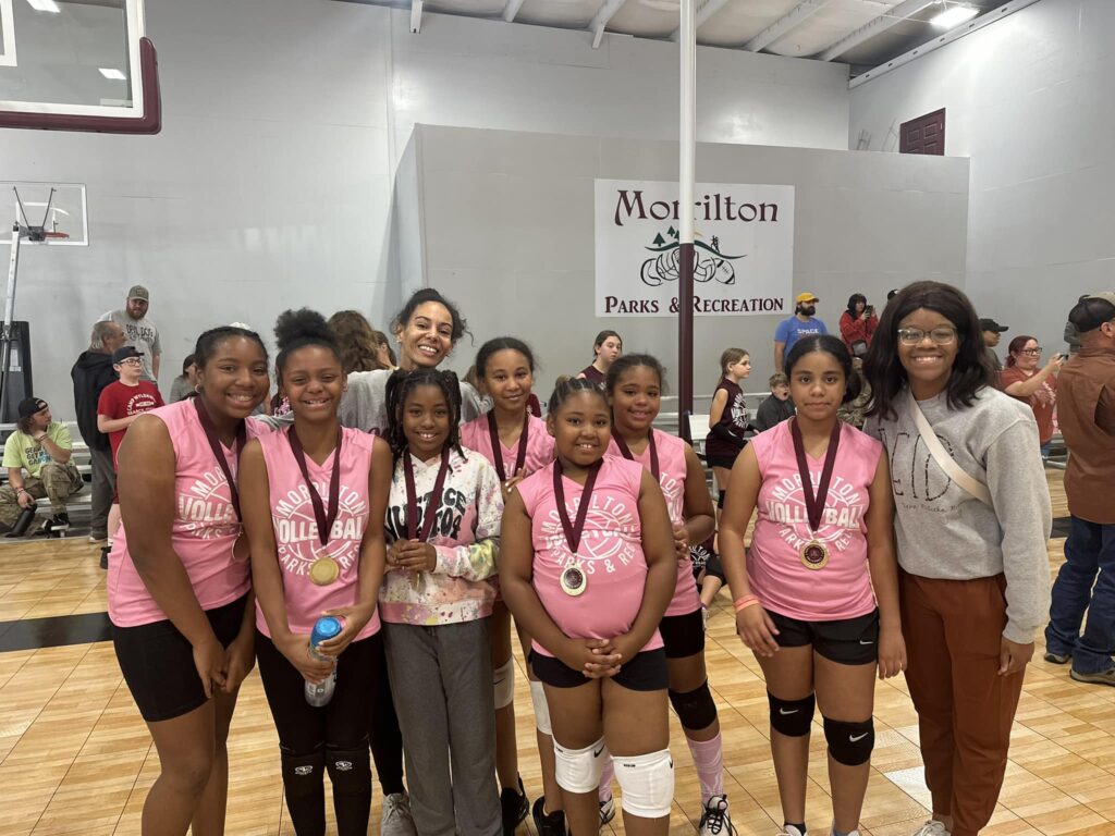 A young girls volleyball team wearing pink tank tops that say "Morrilton Parks and Rec Volleyball." The girls are wearing medals.