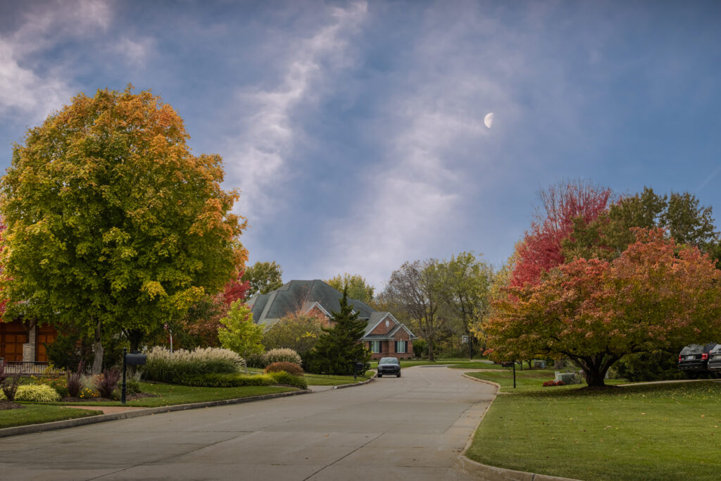 A neighborhood street in the early evening in Autumn. The trees are slowly turning red and yellow and lawns are still green. A half moon is rising in the blue sky.