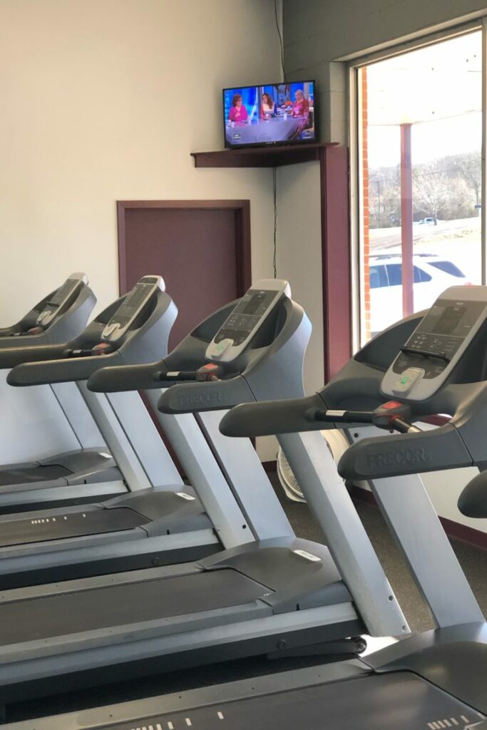 A line of four treadmills in the corner of a gym looking out windows to a parking lot. A small tv is in the upper corner.