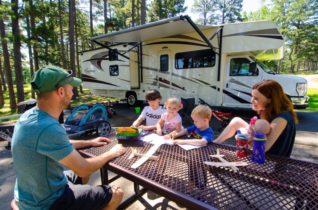 A family of four kids sitting at a brown picnic table beside a Coachmen camper, doing arts and crafts. The boys are picking out crayons and coloring and the little girl is smiling at the camera.