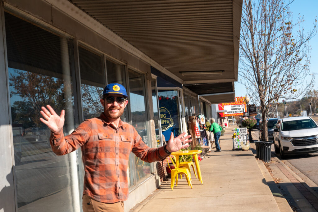 A man waves both hands to the camera as he stands on a sidewalk on a strip of store fronts.