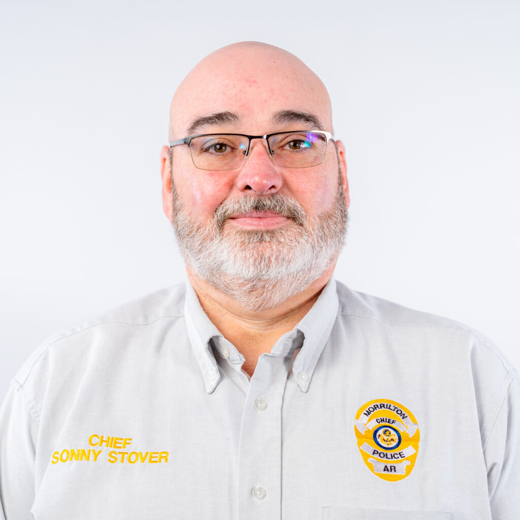 A headshot of Chief of Police in Morrilton, Sonny Stover. Sonny is an older man who is bald with a white thin beard and mustache. He is wearing glasses and a white polo with his badge and title stitched on in yellow.