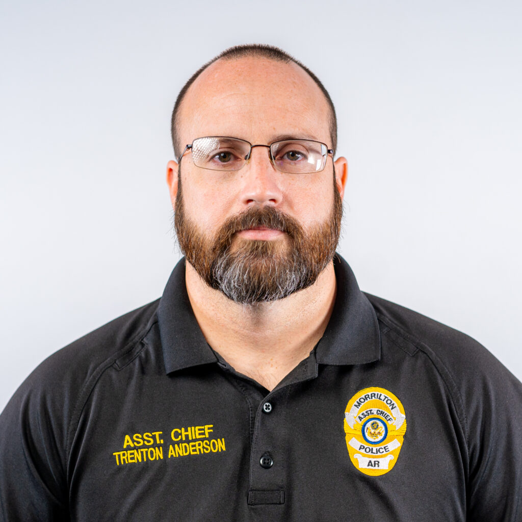 A headshot of the Assistant Chief of Police. He is a white man in his 30s or 40s with a brown beard and mustache and small glasses. He is wearing a black polo with a yellow stitched police badge and his title.